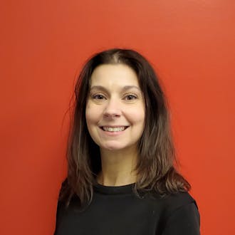 Sarah Koon: a woman with long dark hair smiles. She is wearing a black sweater. The wall behind her is red.