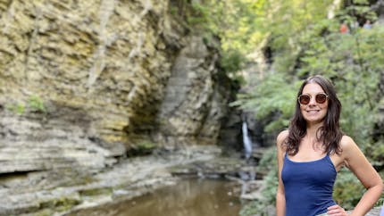 Sarah Koon wears sunglasses, a blue tank top, and green shorts on the right side of this photo. Lush green trees are behind her, and rock walls line a creek on the left side of the photo, with a waterfall toward the center. 