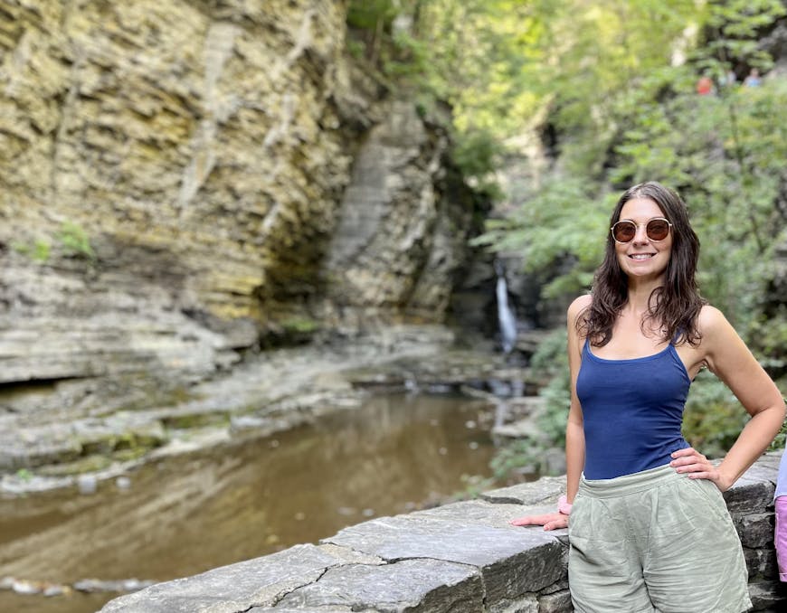 Sarah Koon wears sunglasses, a blue tank top, and green shorts on the right side of this photo. Lush green trees are behind her, and rock walls line a creek on the left side of the photo, with a waterfall toward the center.