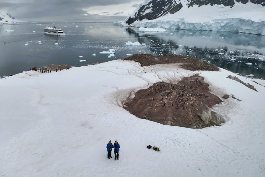 Oceanites researchers at Neko Harbour, Antarctica, monitoring a Gentoo penguin colony with snow and cloudy skies. A ship is in the top left quadrant of the photo. The photo was taken by a drone. 