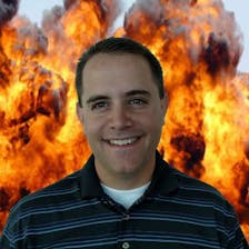 A man smiles in front of a fiery scene. 