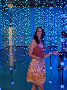 Sarah Koon stands in a room with strands of spherical lights, smiling.