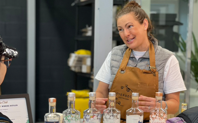 A woman in an apron engages with a group of guests with bottles of gin lined up in front of her
