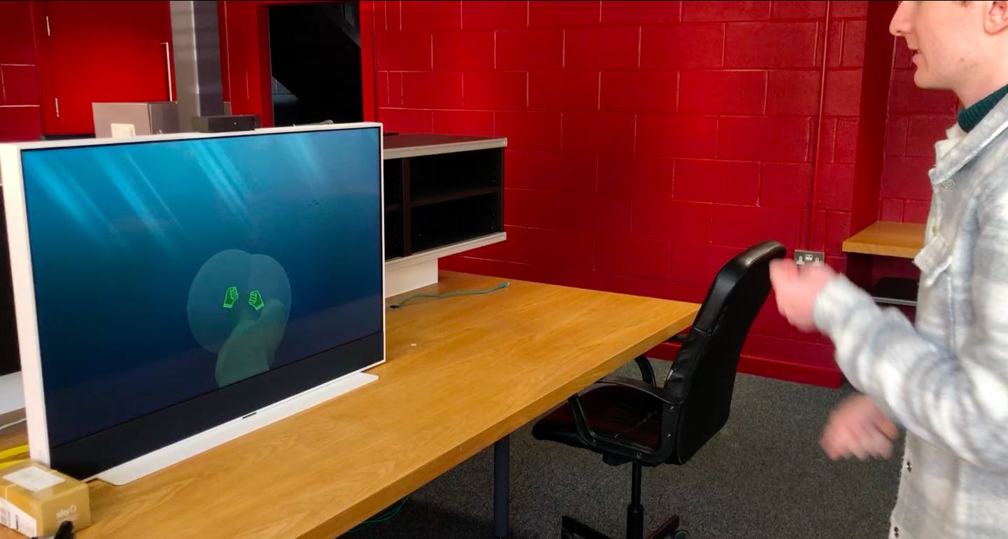 A large TV screen sitting on a desk, in an office with red walls. Someone is playing an interactive, gesture-based experience on the TV screen, pulling themselves through water along a rope.