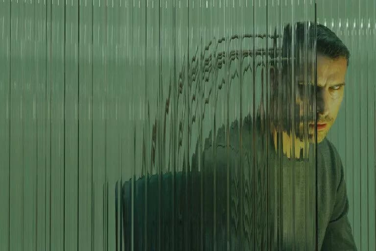 A man with short dark hair and a grey top, sitting on a black chair. He is viewed through a pane of distorted glass. Thick wires appear to be connected from the chair into the back of his head. His eyes are half closed. 