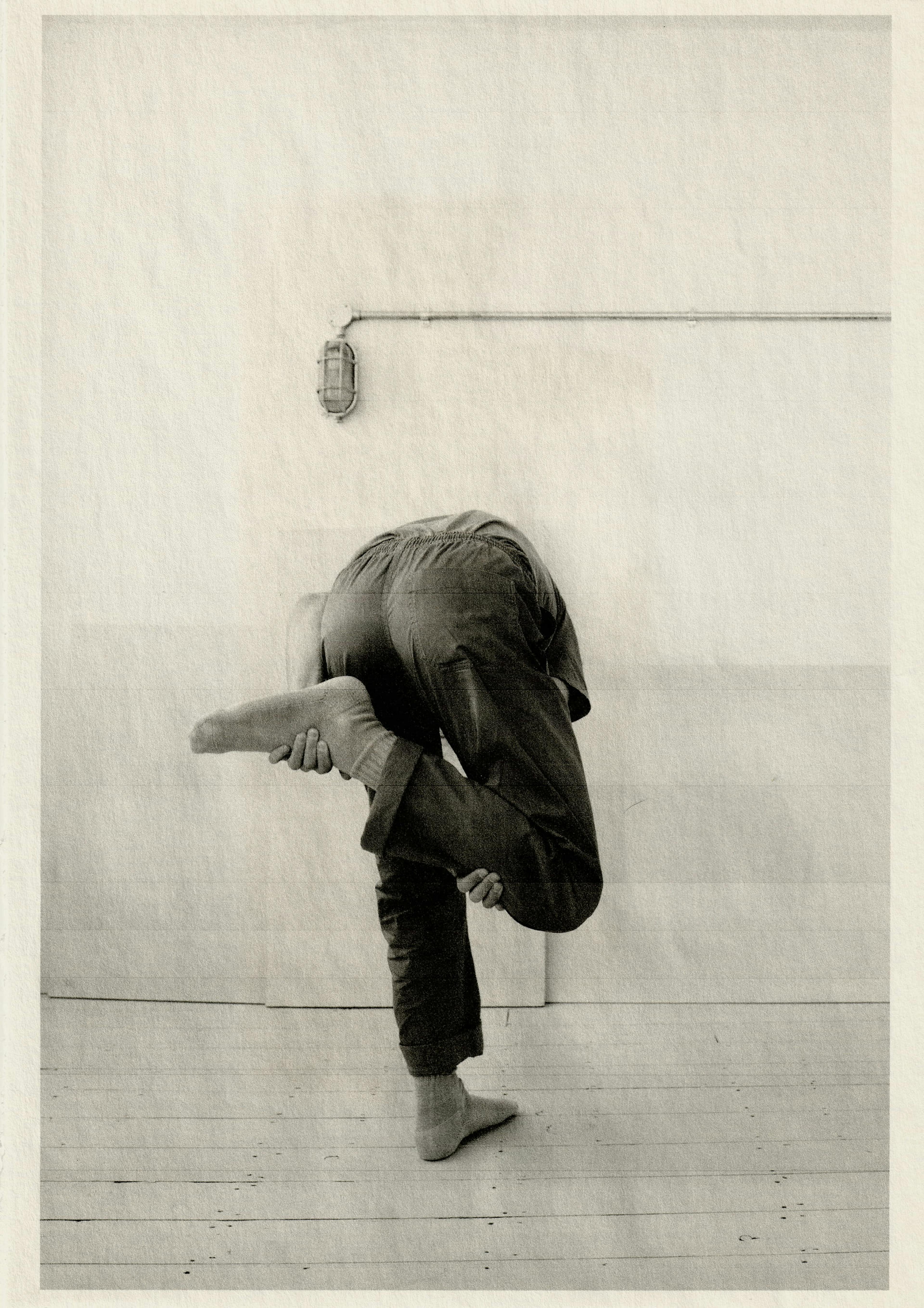 Black and white photograph of a human figure standing on his left leg while holding the right leg with his back turned towards the camera and his head concealed.