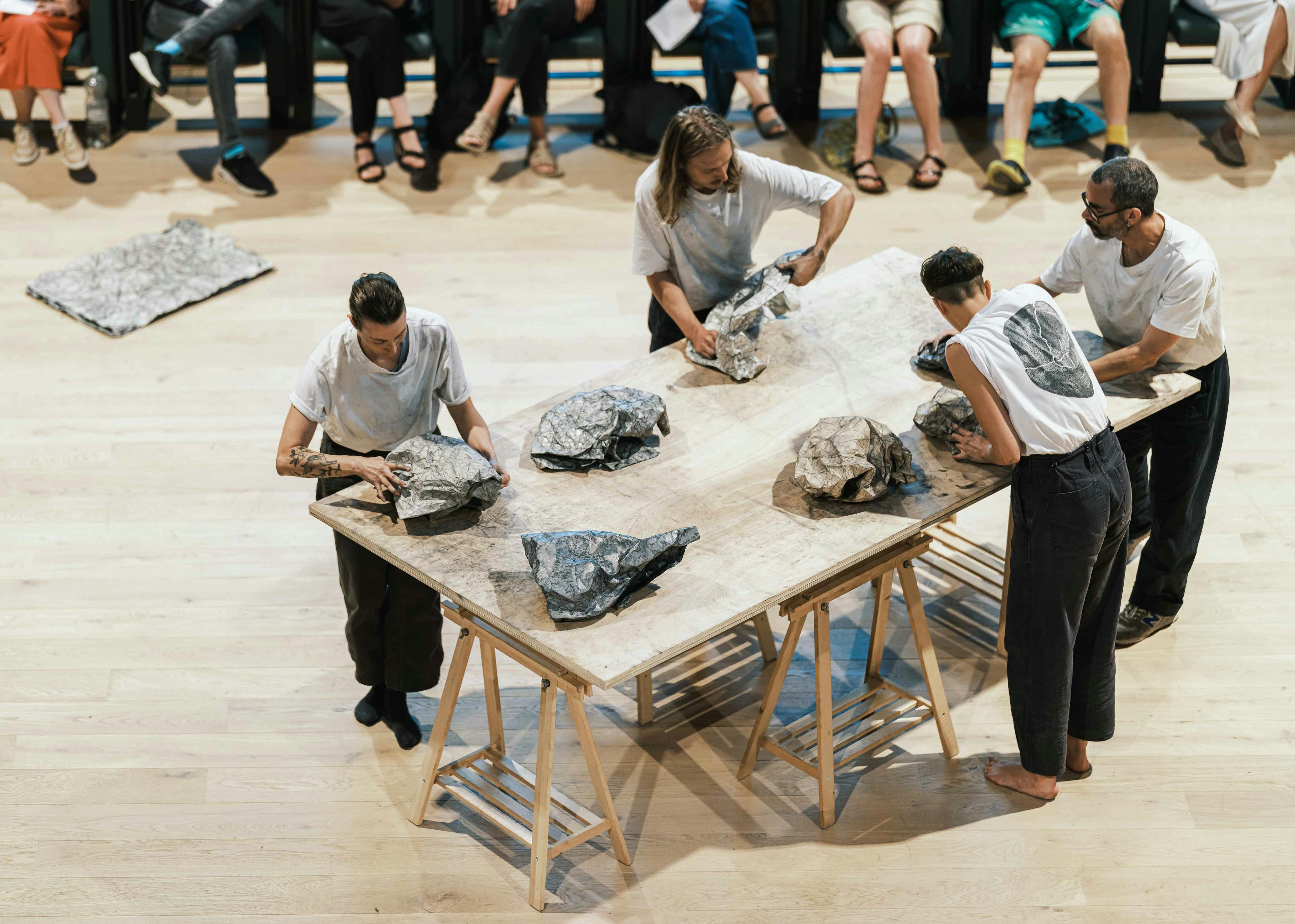 Photograph of four performers wearing grey and white clothing standing around a table constructing stone shapes from graphite covered paper. 