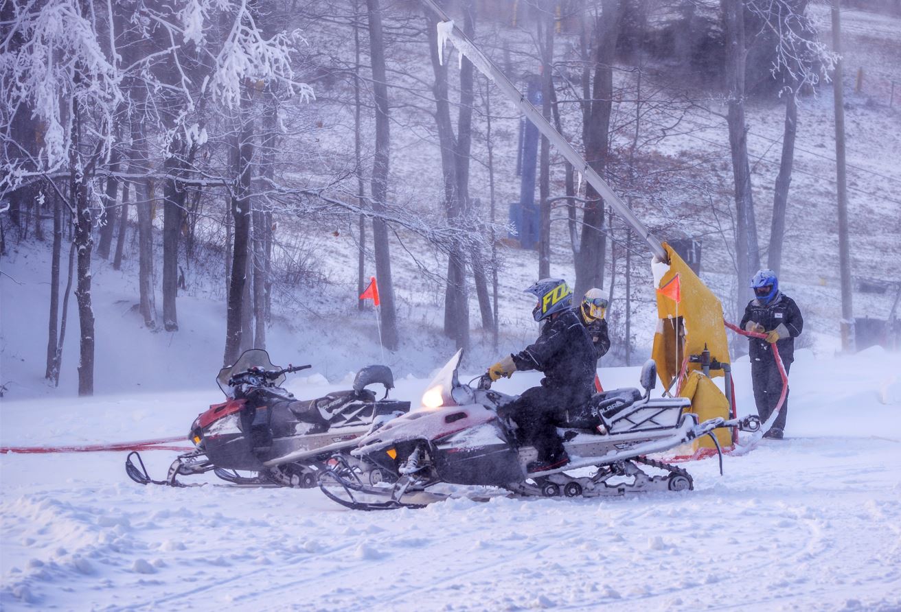 Three Snowmakers working at Chicopee. One sitting on a snowmobile, while two check hoses attached to a snow gun. 