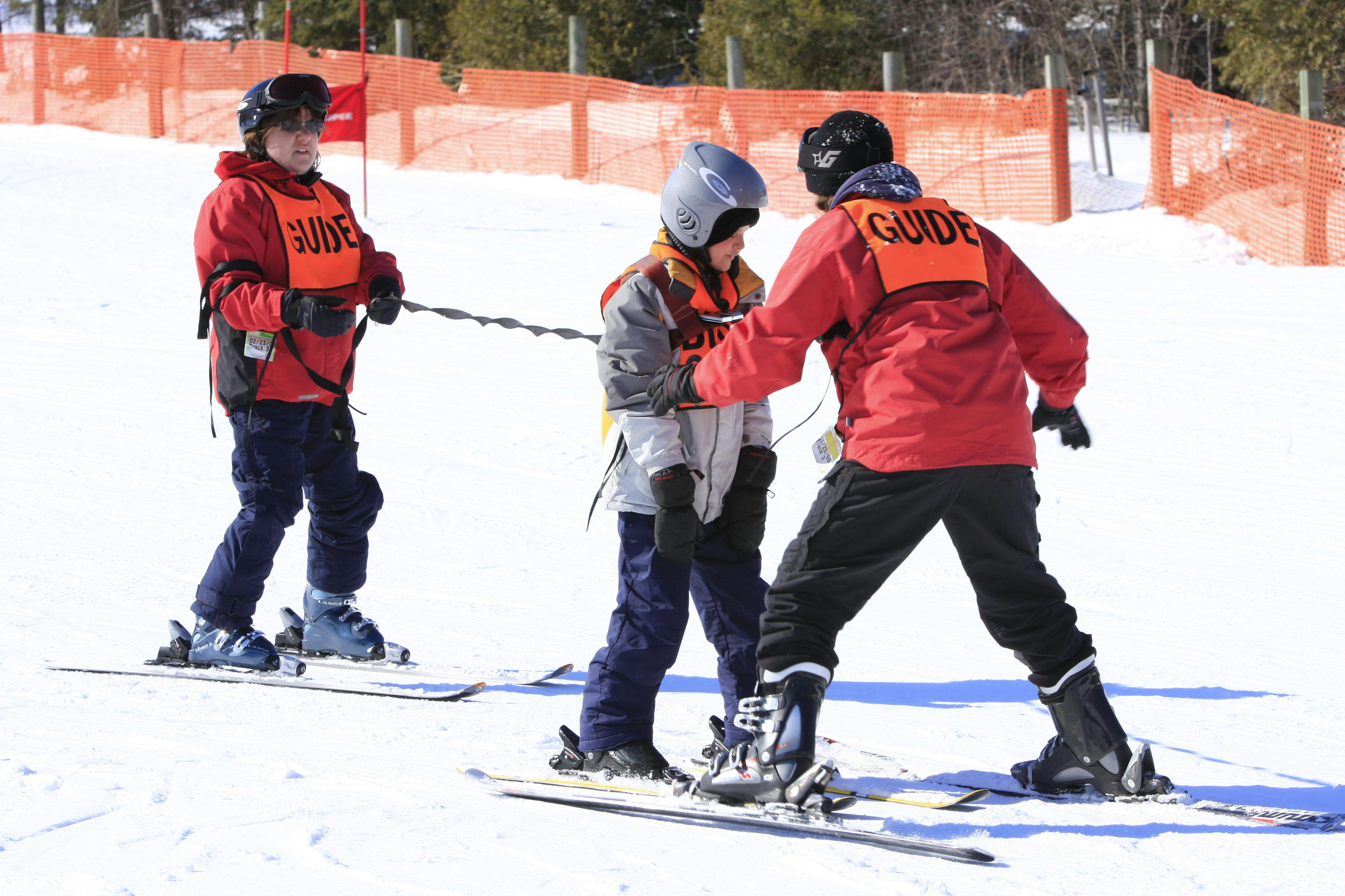 Track-3 student in a harness and tether with 2 guides assisting them. All 3 people are in skis. 