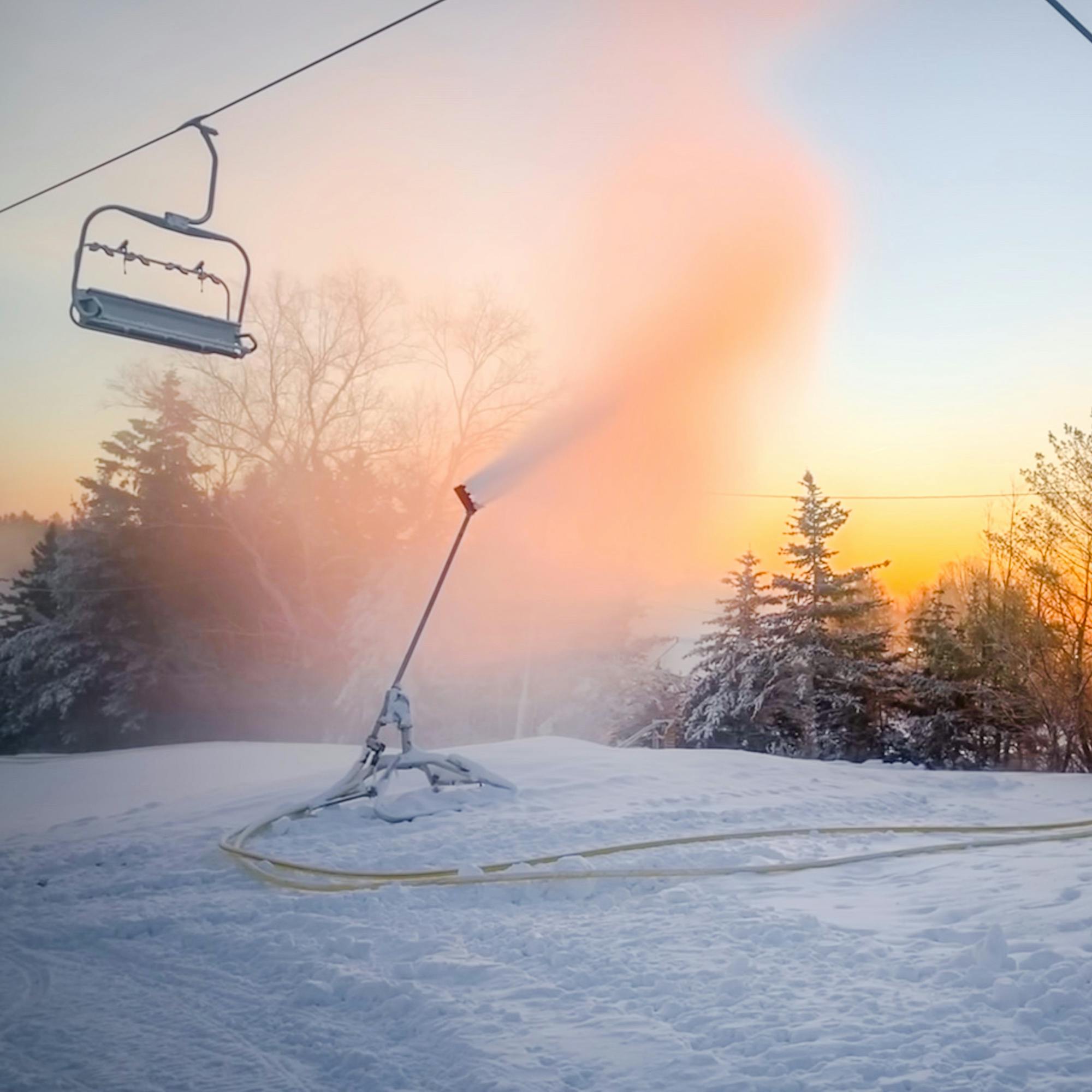 Scenery shot: Early morning sun rising behind the trees. A snow gun blowing snow in the center, and an empty chairlift chair in the top left. 