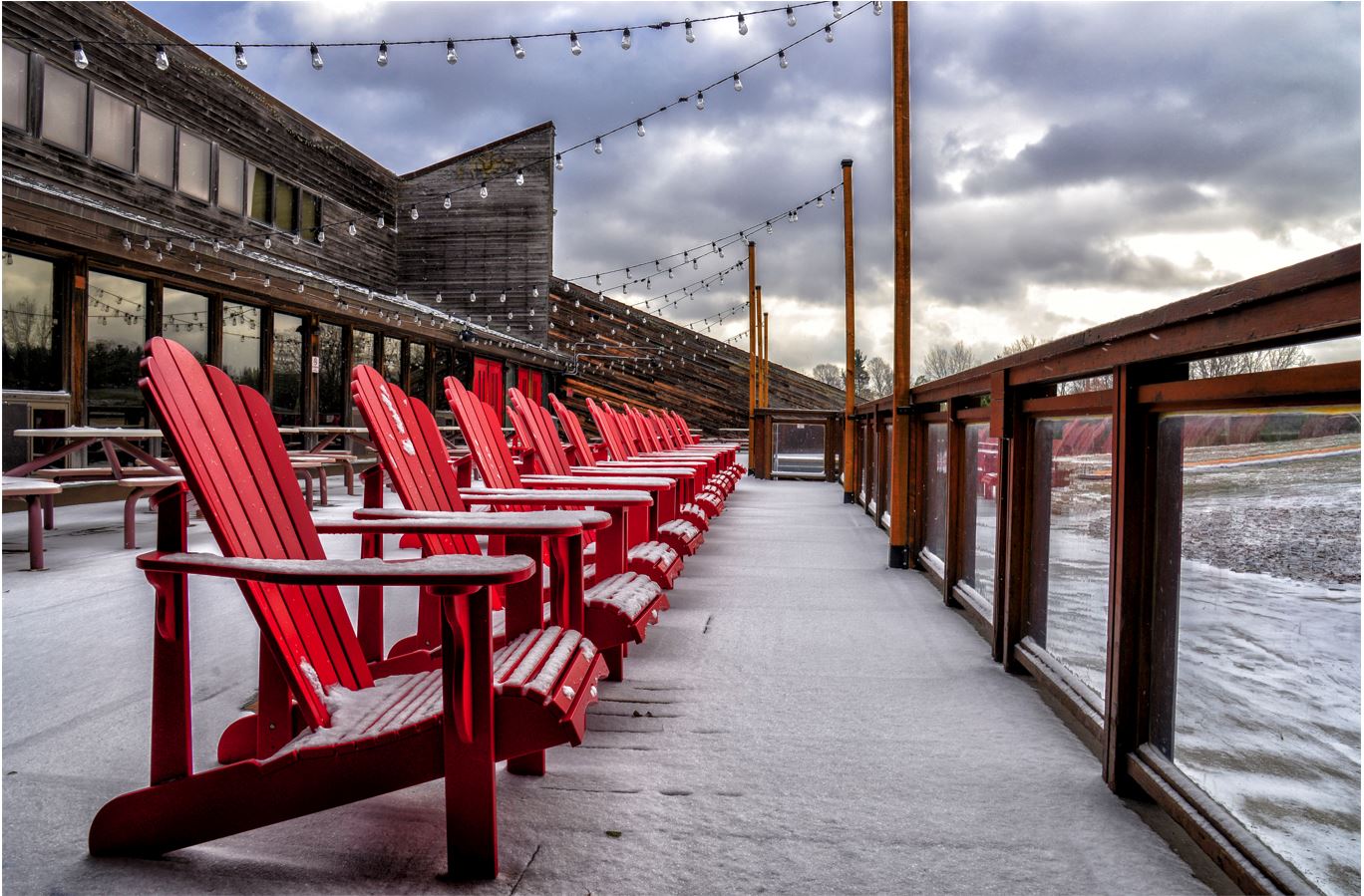 A line of red Adirondack chairs on a snowy deck. 