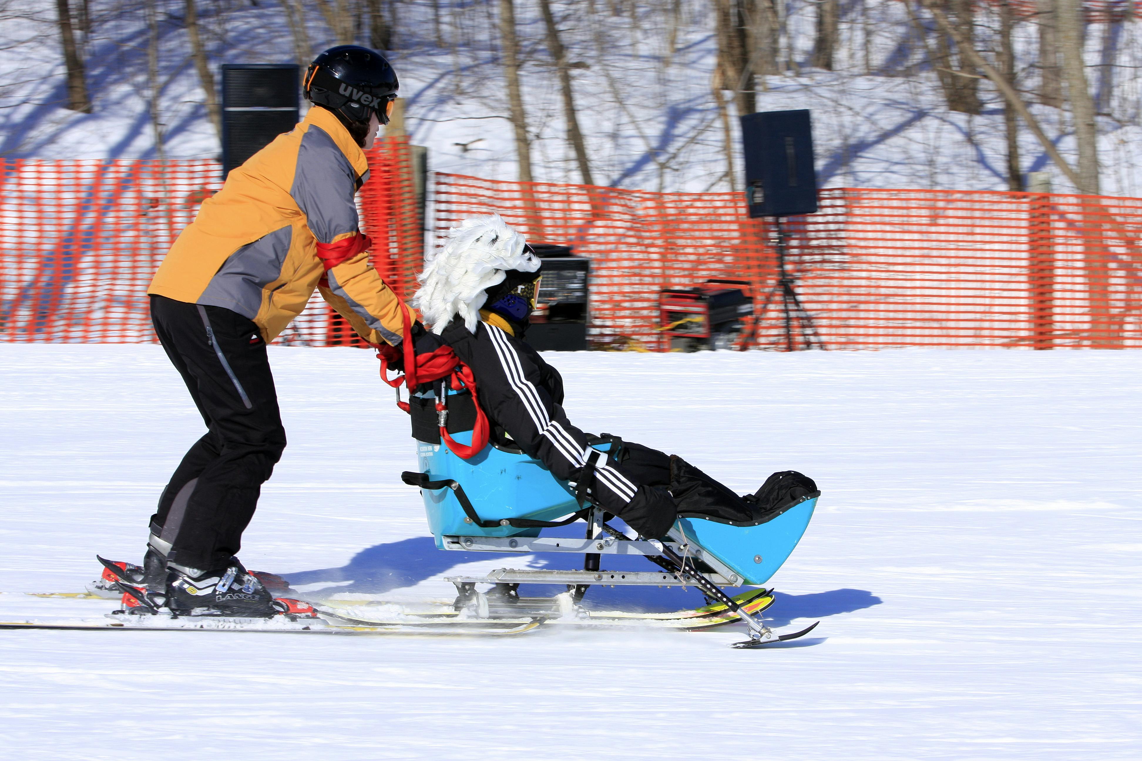 Track-3 volunteer holding the back of a sit ski with a student in it, skiing down the hill. 