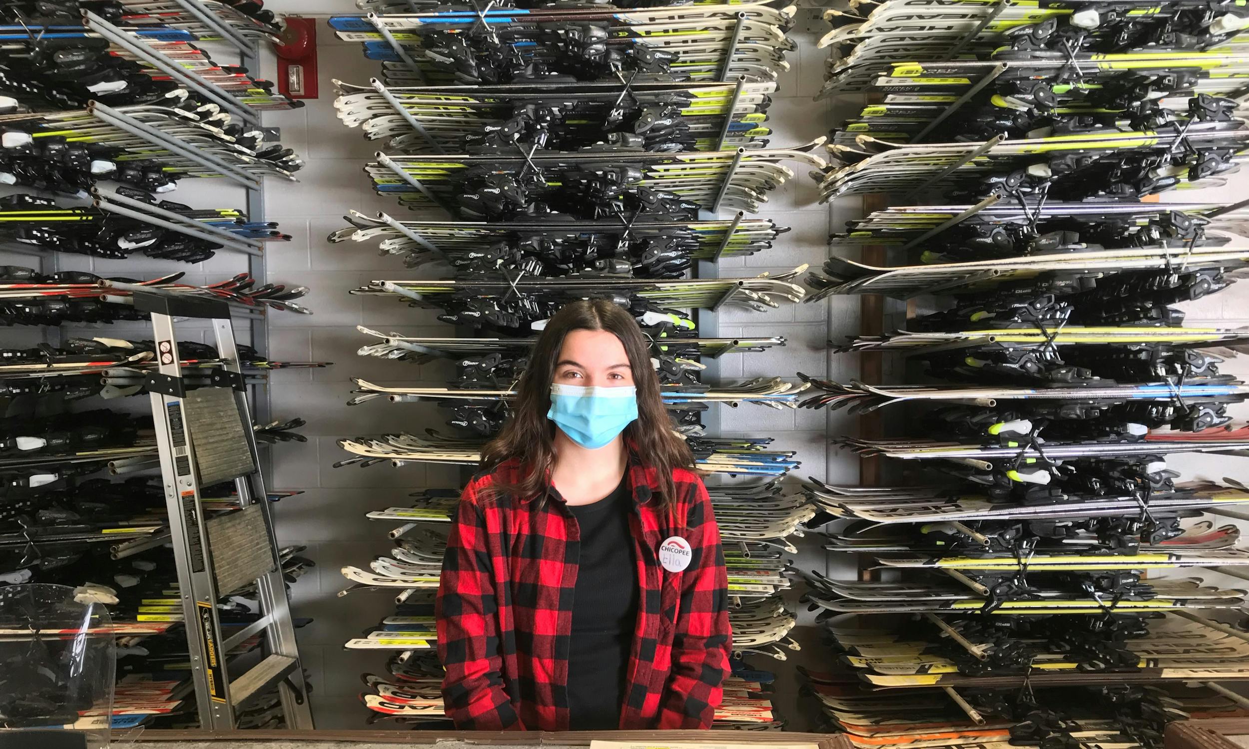 Rental Shop Attendant Ella standing in front of racks of rental skis, her smile hidden by her mask. 
