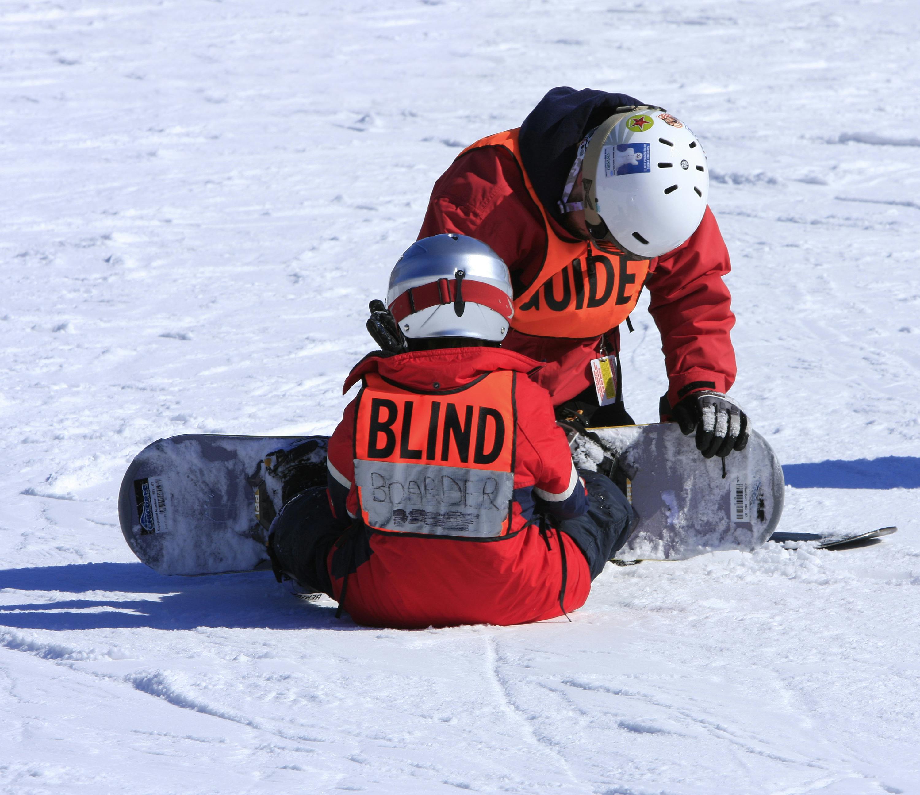 A snowboarder wearing a "Blind Boarder" vest and their guide (wearing a "Guide" vest), sitting on the hill adjusting bindings.