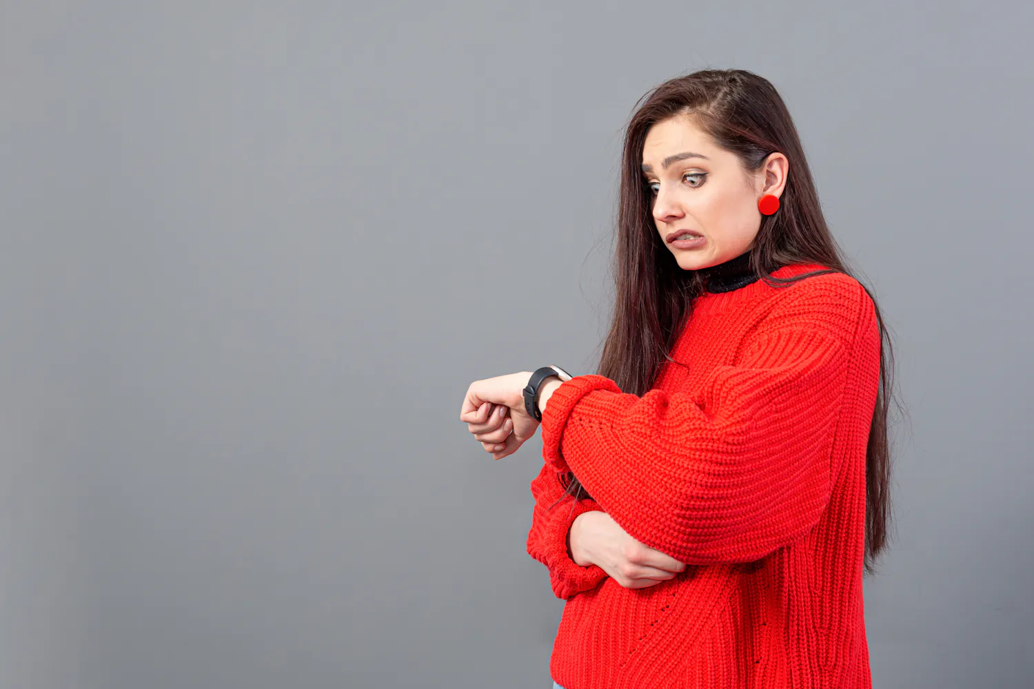 Femme vêtue d'un pull rouge regardant sa montre sur fond gris.