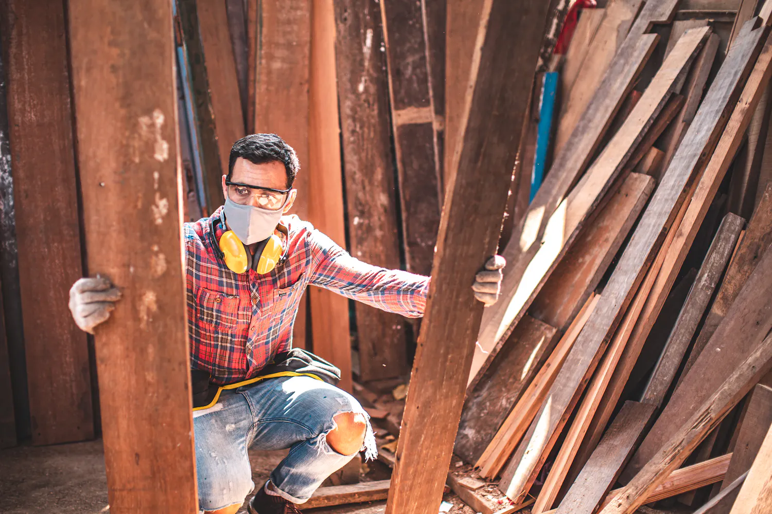 Homme en tenue de travail tenant deux grandes planches en bois au milieu d'autres planches.