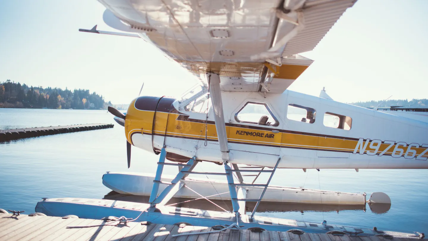 Kenmore Air seaplane docked on floats, yellow and white livery, on the water near a wooded shoreline