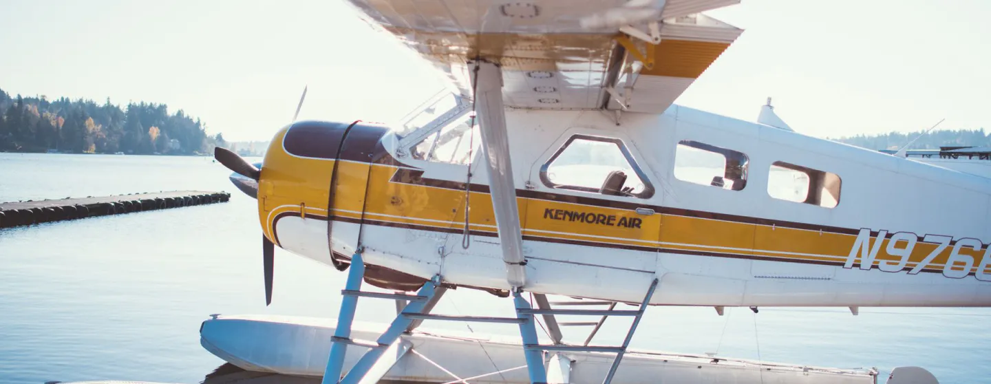 Kenmore Air seaplane docked on floats, yellow and white livery, on the water near a wooded shoreline