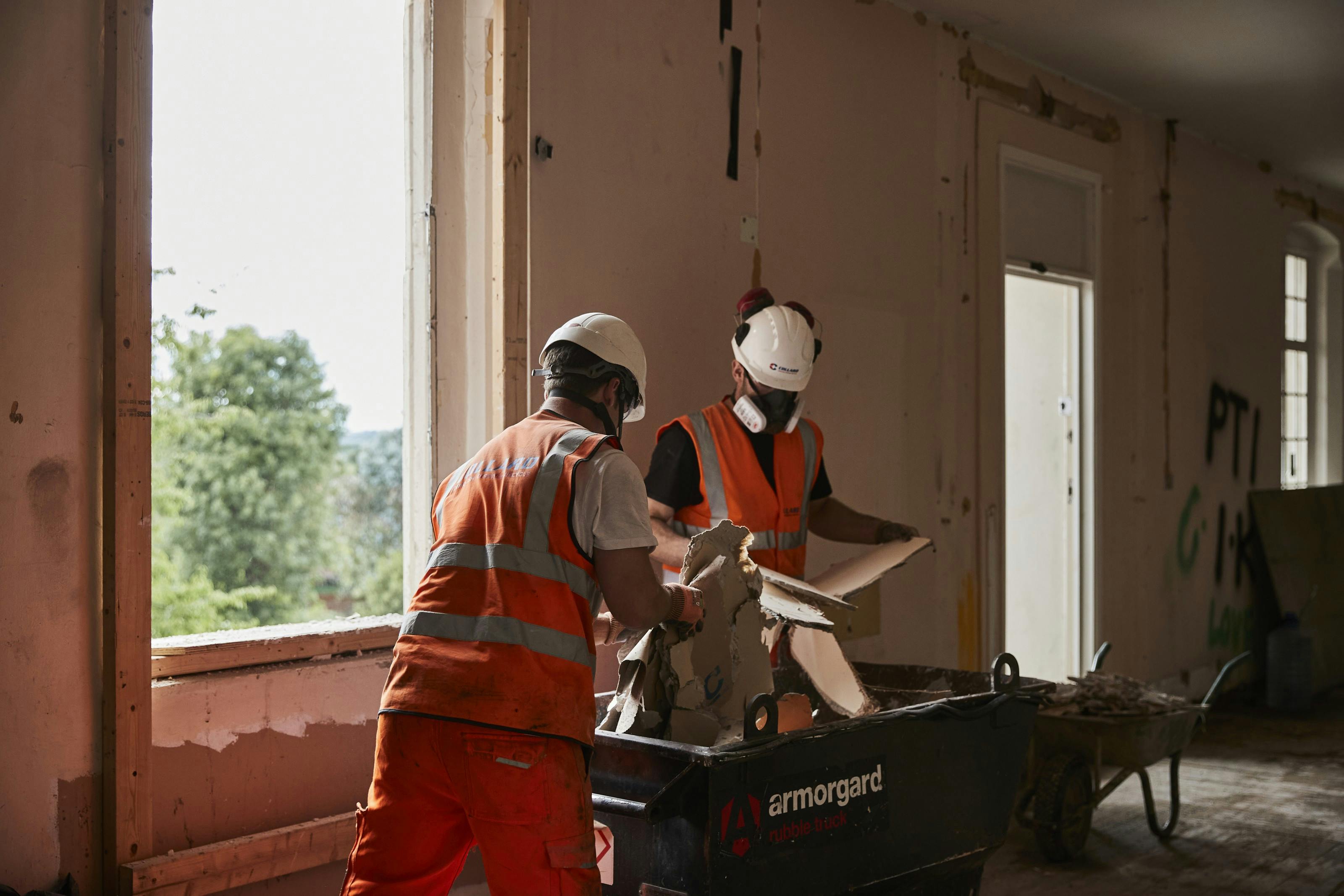 Two workmen disposing of waste into a small skip on a renovation project.