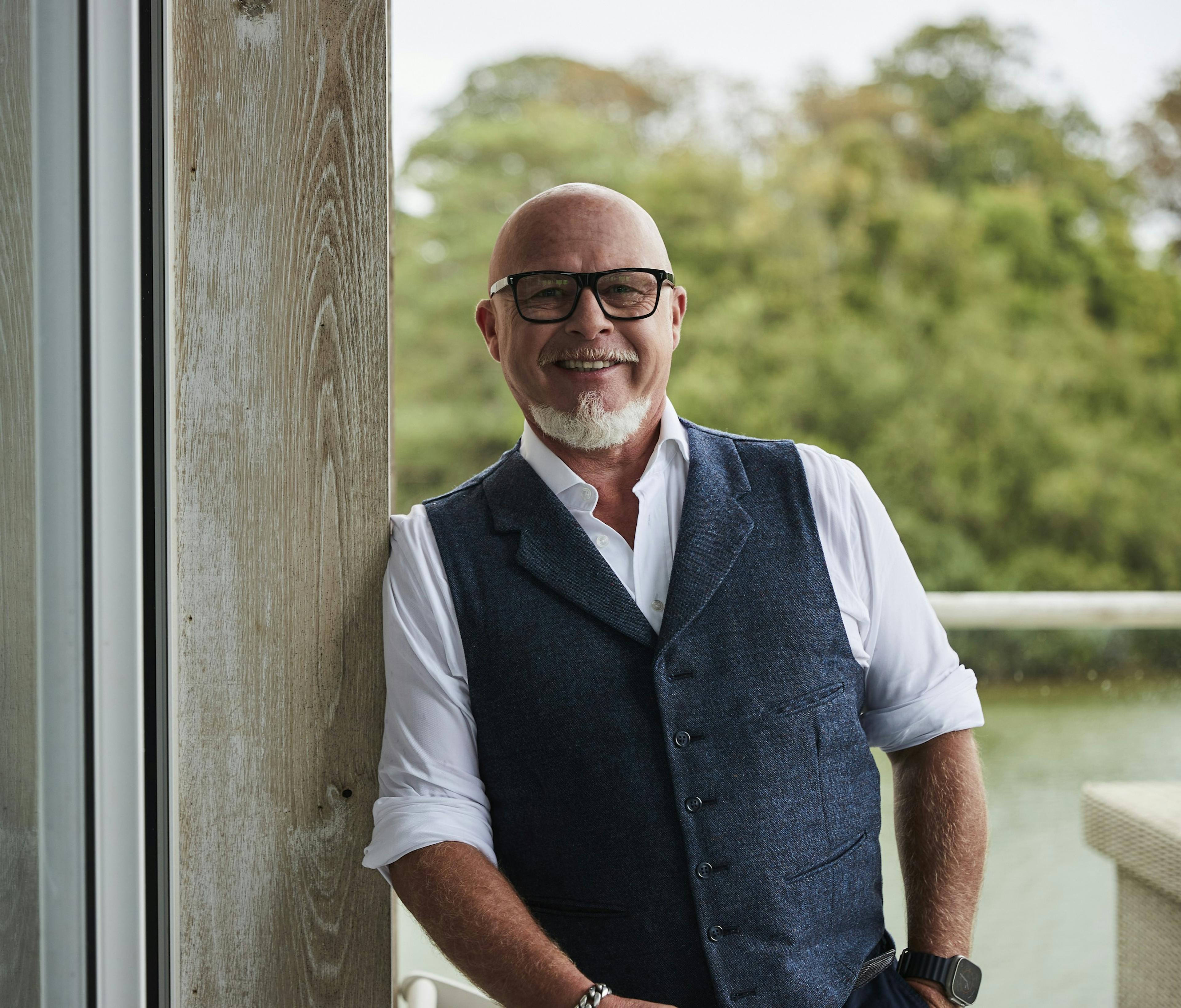 A man in glasses, shirt and waistcoat leaning against a wall