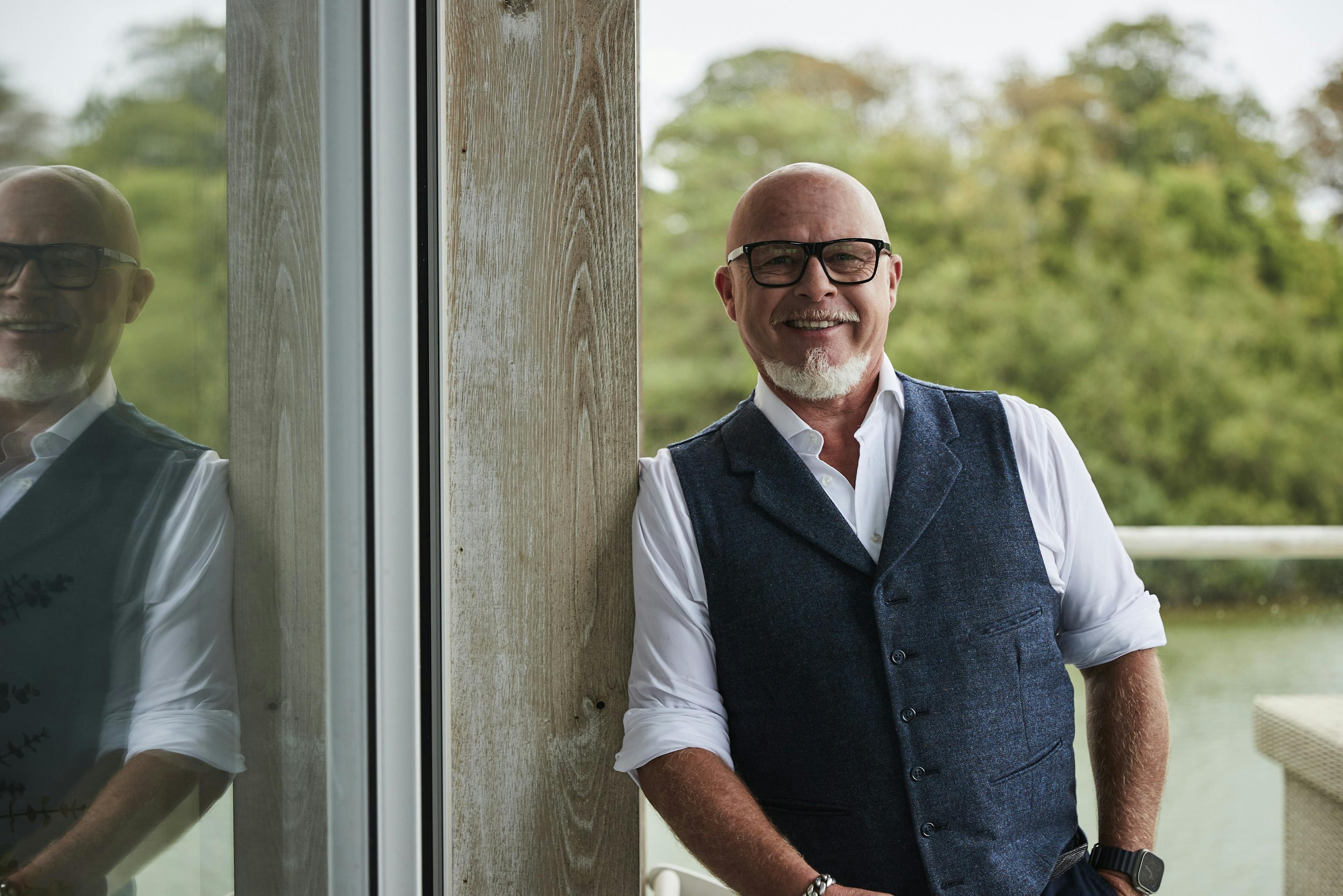 A man in glasses, shirt and waistcoat leaning against a wall