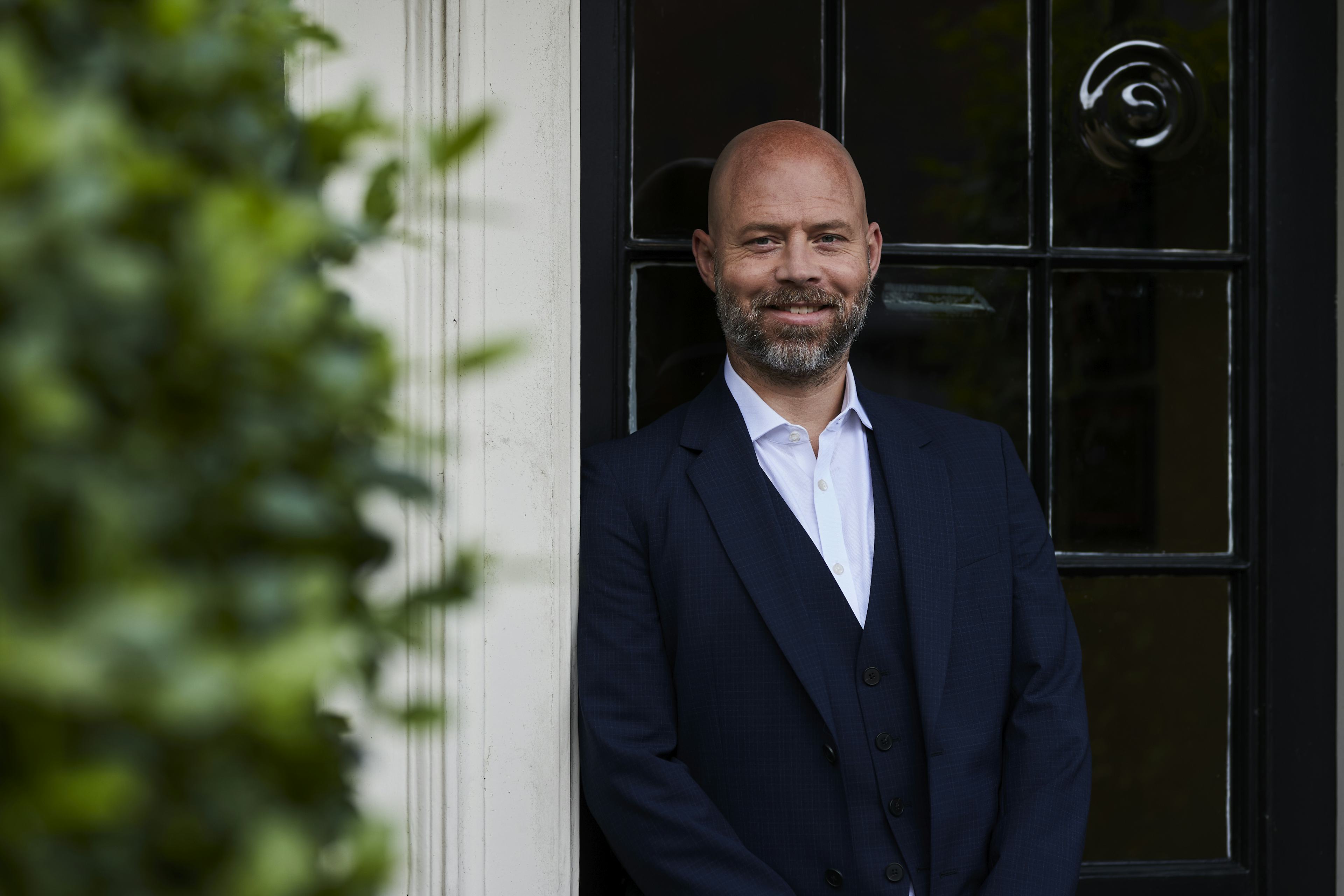 Caption: a bald man in a suit standing in front of a door