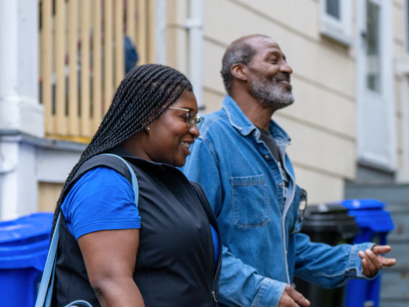 Image of a smiling community health partner walking alongside a middle-aged man wearing a denim jacket outdoors in a neighborhood.