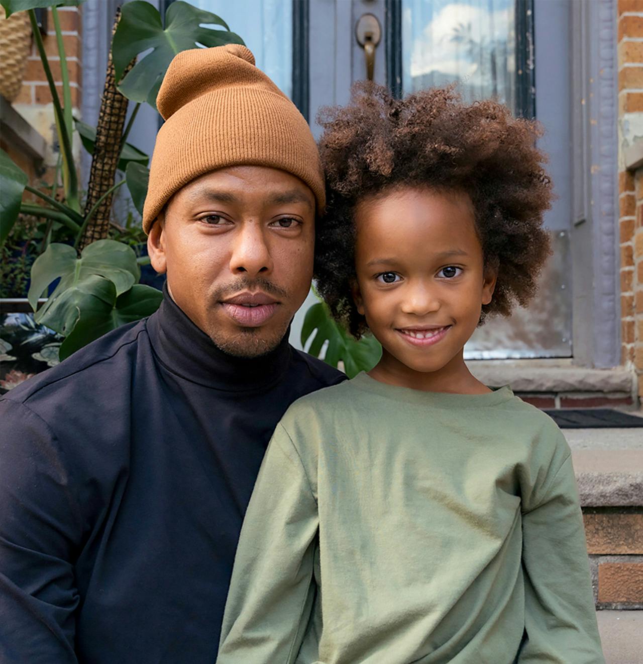 Person with a child wearing a black turtleneck and green shirt sits on stoop stairs smiling.
