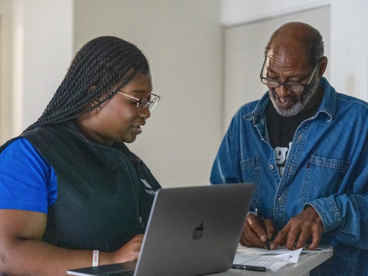 Image of a community health partner showing a laptop to an older man wearing glasses as he signs paper documents.