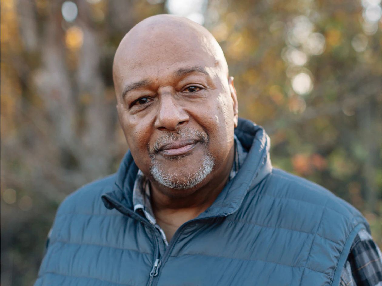 Image of an older member with a goatee wearing a blue vest, standing outdoors with trees in the background.