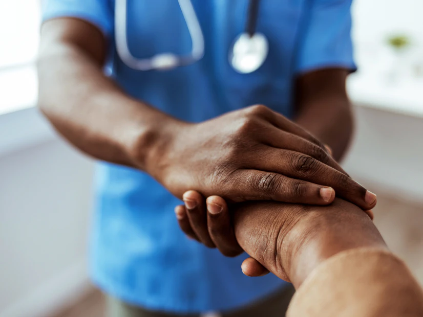 Image of a healthcare worker in blue scrubs gently holding an older patient's hand in a reassuring, supportive gesture.