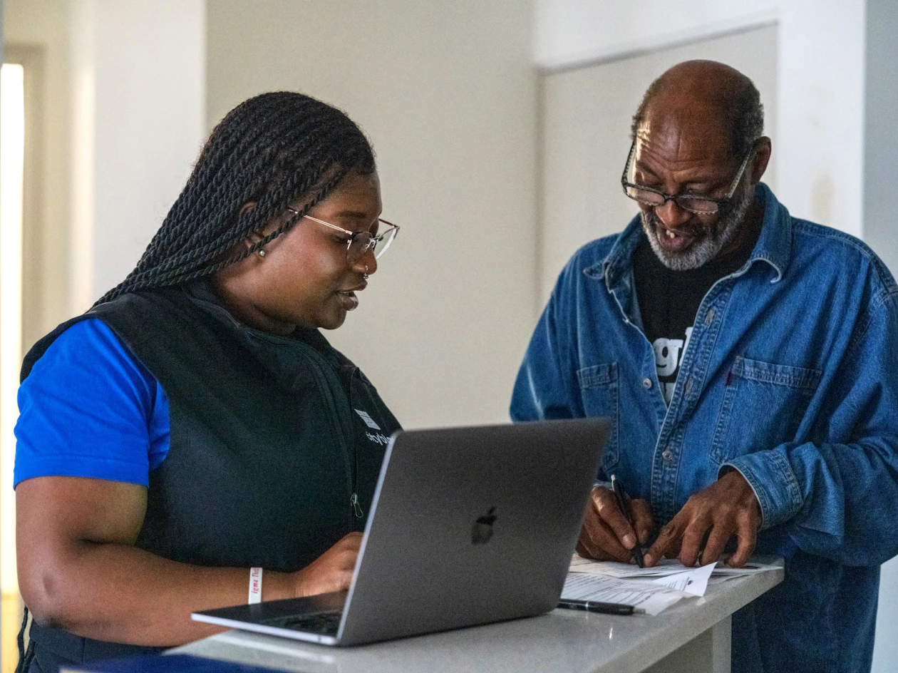 Image of a community health partner showing a laptop to an older man wearing glasses as he signs paper documents.