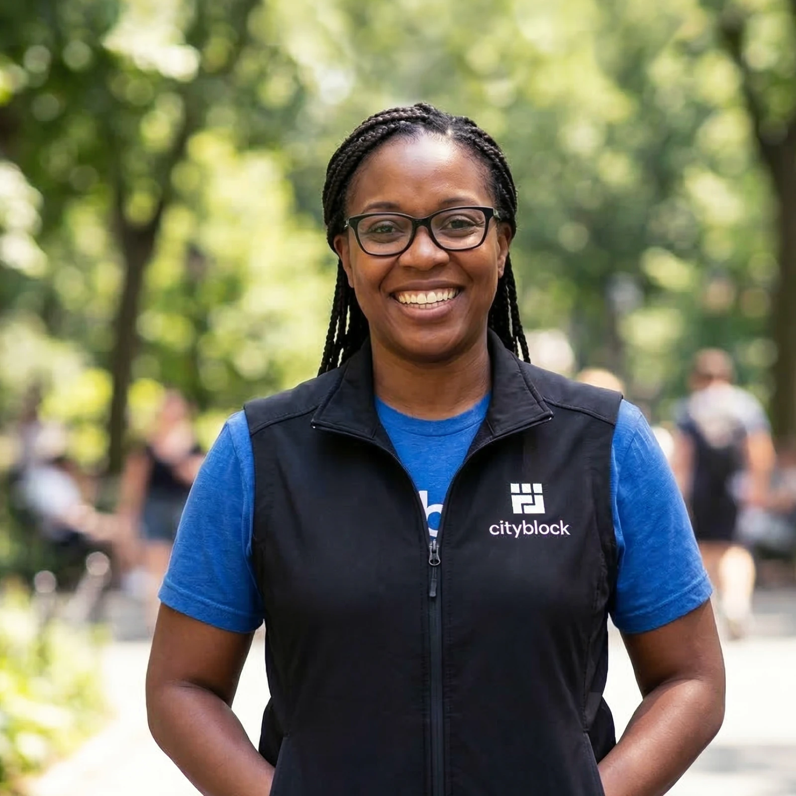 Image of a smiling community health partner with braided hair wearing glasses and a Cityblock vest standing outdoors on a tree-lined walkway.