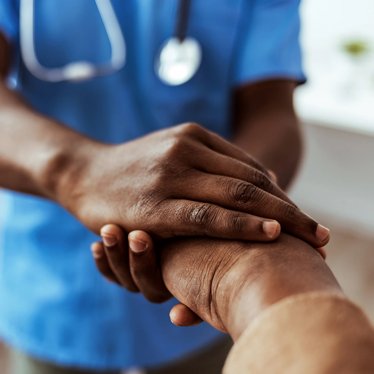 Image of a healthcare worker in blue scrubs gently holding an older patient's hand in a reassuring, supportive gesture.