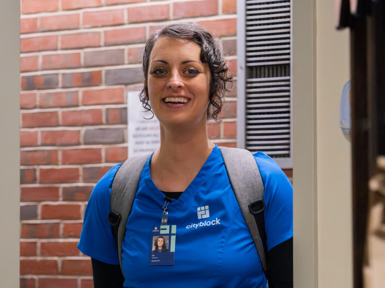 Image of a smiling nurse in blue scrubs with a backpack standing in front of a red brick wall.