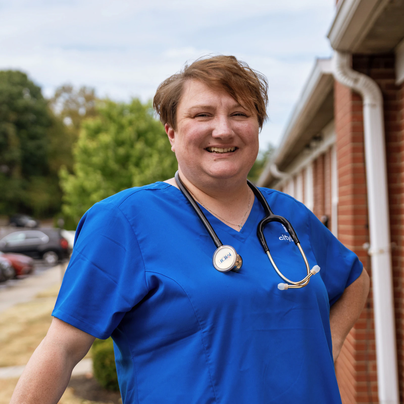 Image of a healthcare provider wearing blue scrubs and a stethoscope, smiling outside a brick building.