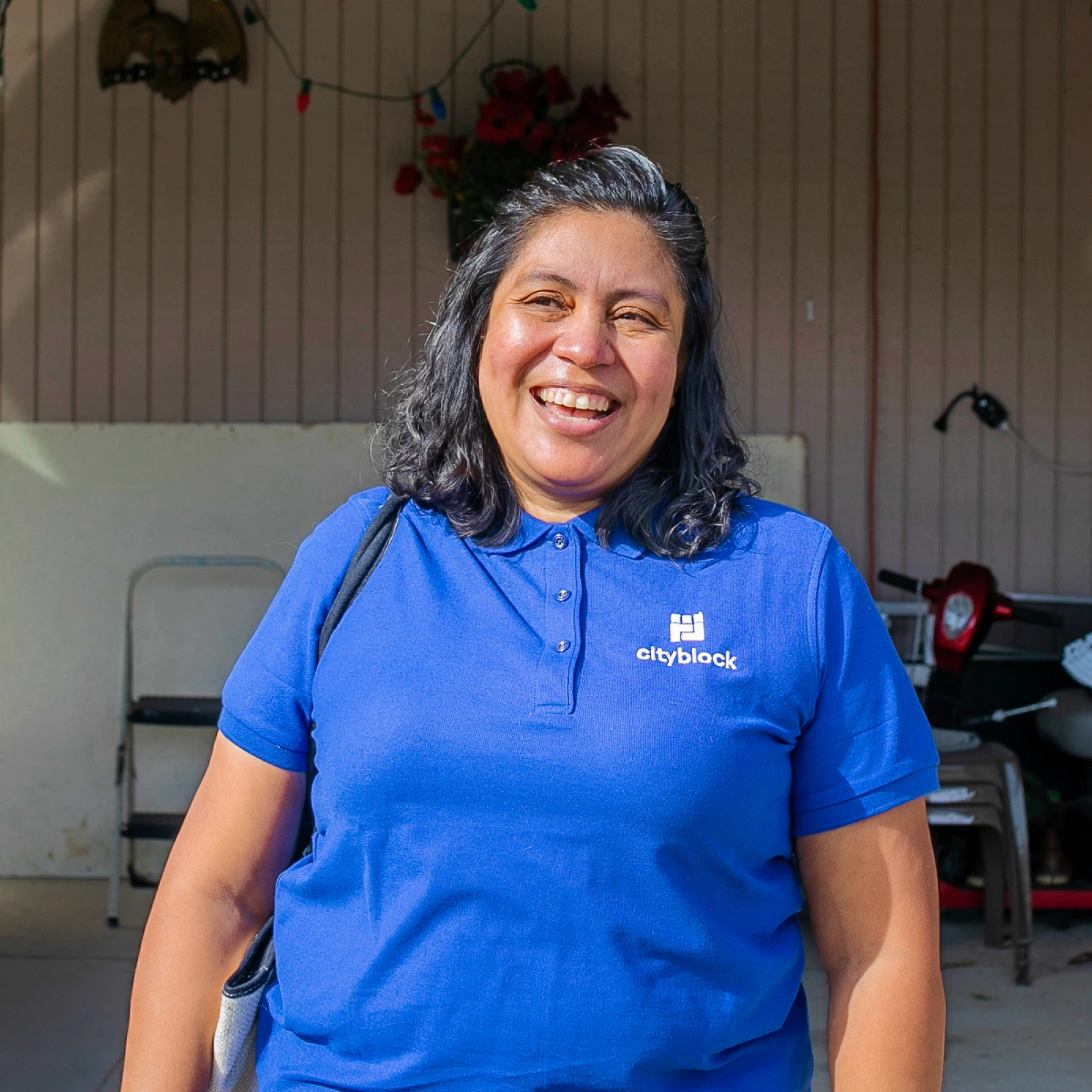 Image of a community health partner wearing a blue Cityblock polo shirt standing and smiling under a covered driveway outside a home.