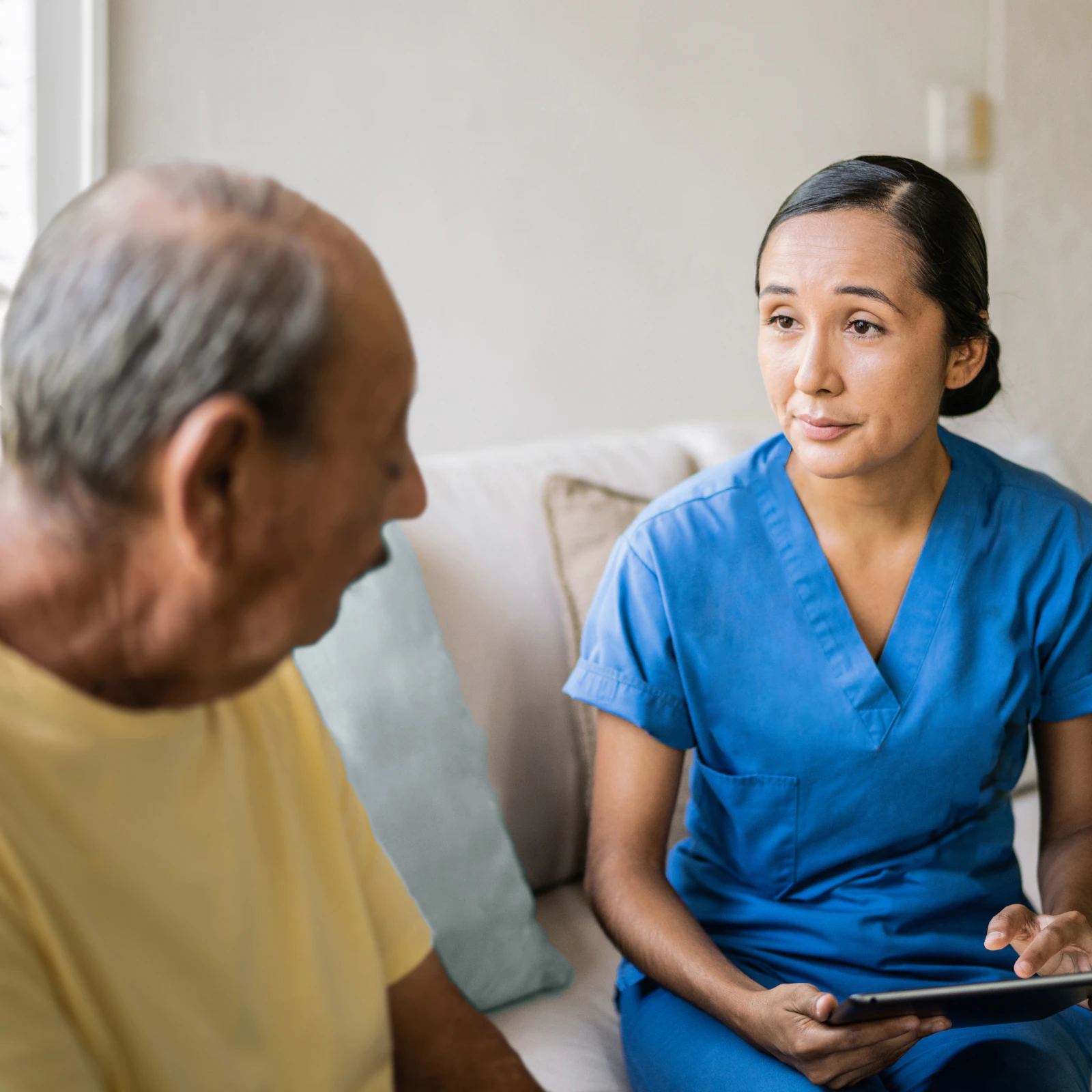 Image of a healthcare worker in blue scrubs holding a tablet while sitting on a couch and conversing with a senior member.