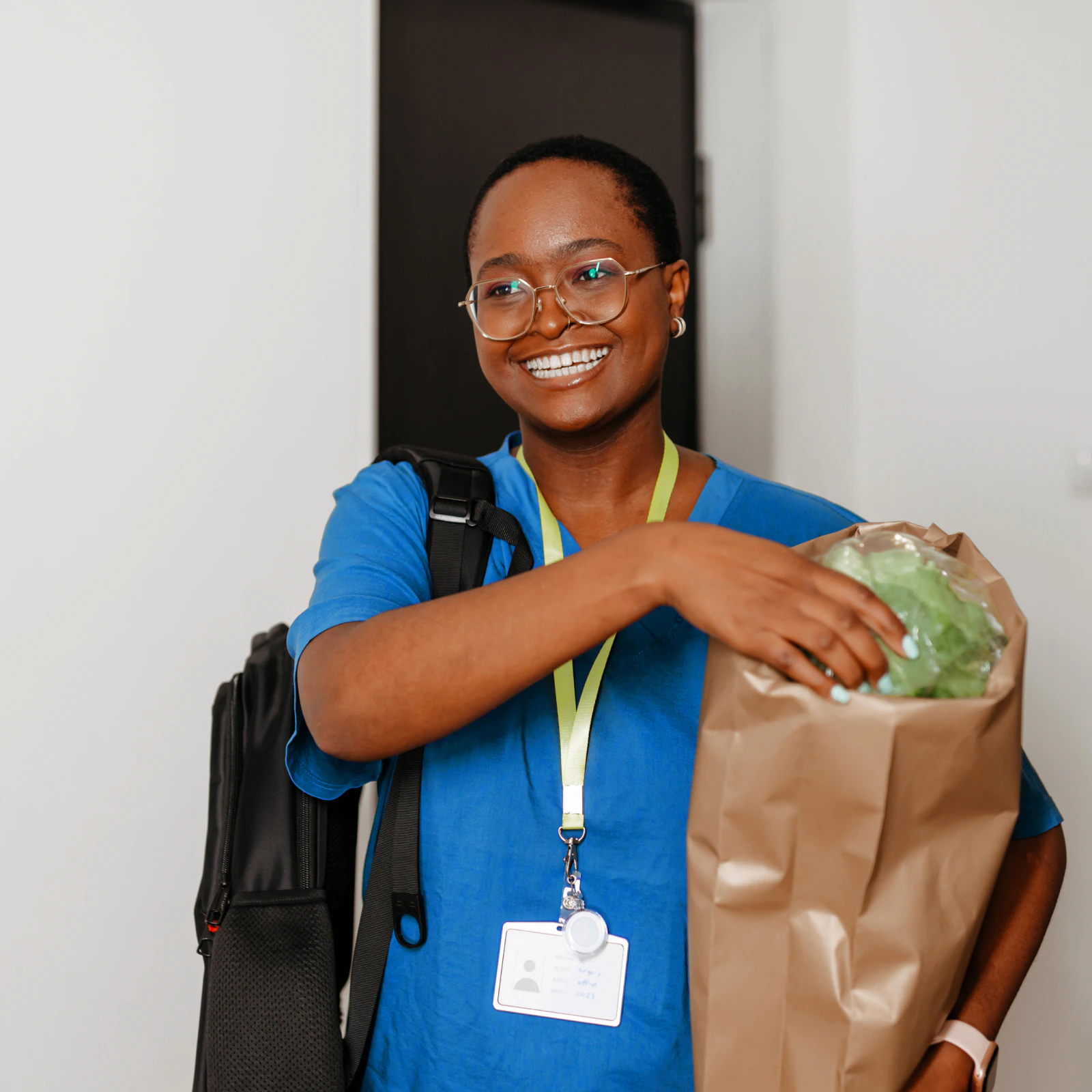 Image of a healthcare worker in blue scrubs holding a brown paper bag of fresh produce while wearing a backpack.