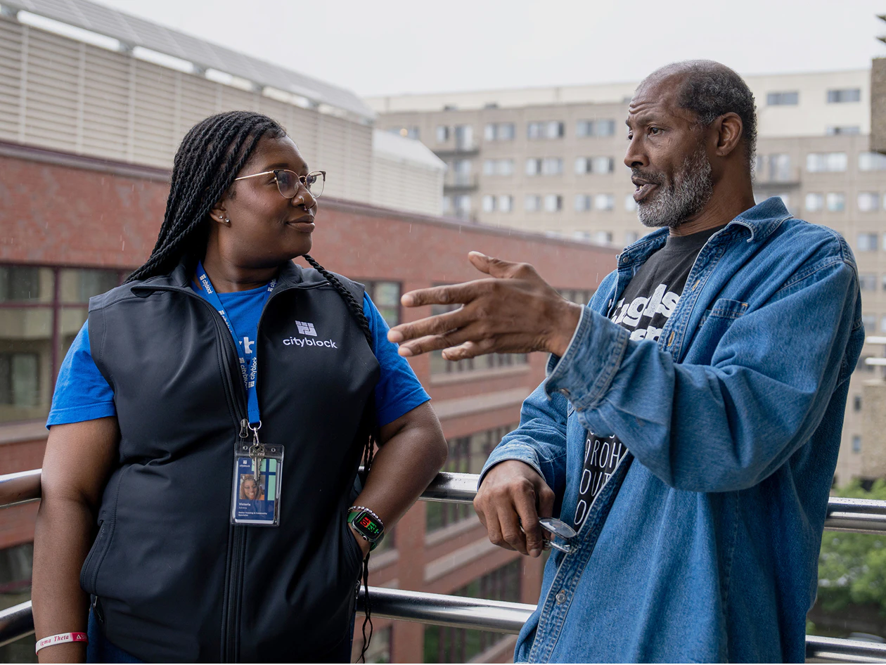Image of a community health partner wearing a Cityblock vest standing on a balcony listening to a man in a denim jacket gesture while speaking.