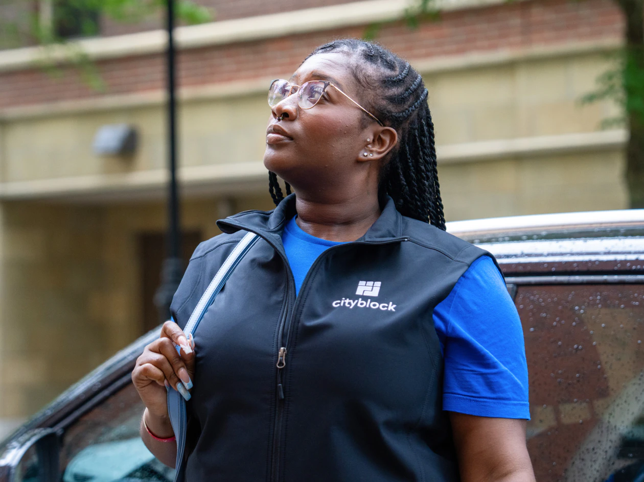 Image of a community health partner with braided hair and glasses wearing a Cityblock vest, looking upward while standing outdoors near a vehicle.