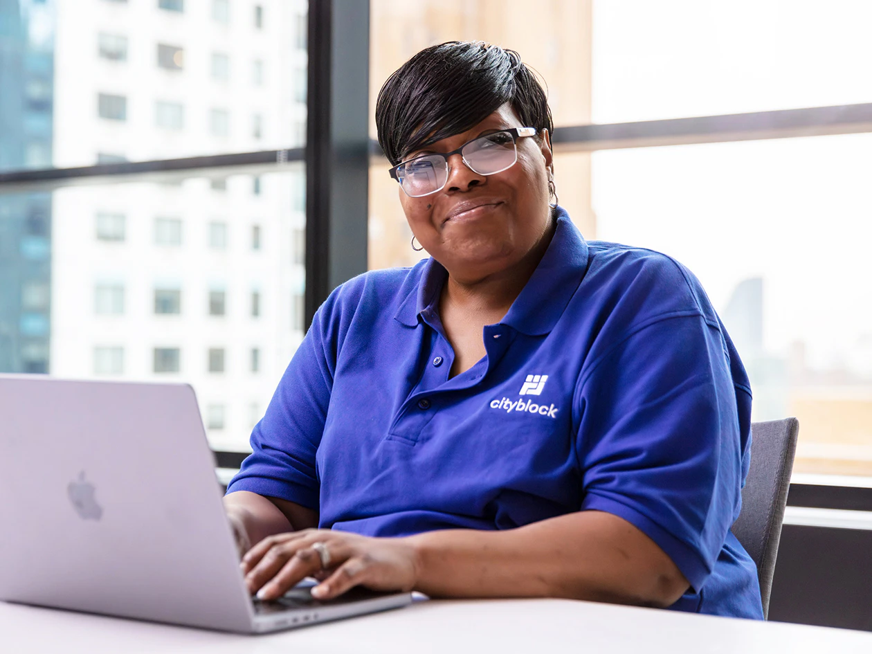 Image of a community health partner wearing glasses and a blue Cityblock polo shirt smiling while working on a laptop at a table.