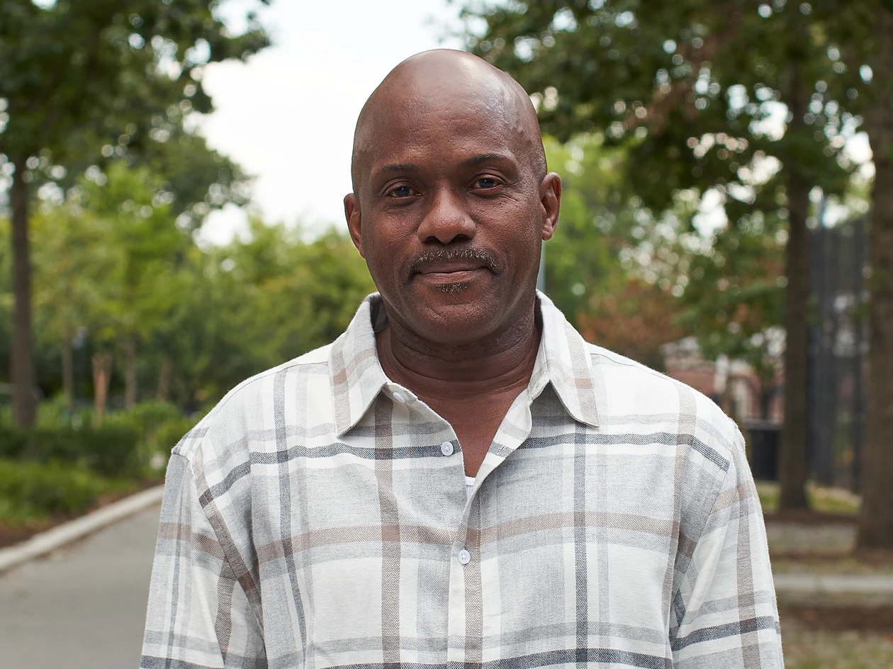 Image of a middle-aged man wearing a plaid button-down shirt standing outdoors with green trees in the background.