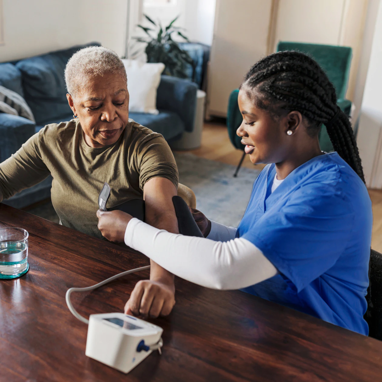 Image of a healthcare worker in blue scrubs placing a blood pressure cuff on an older woman sitting at a table.