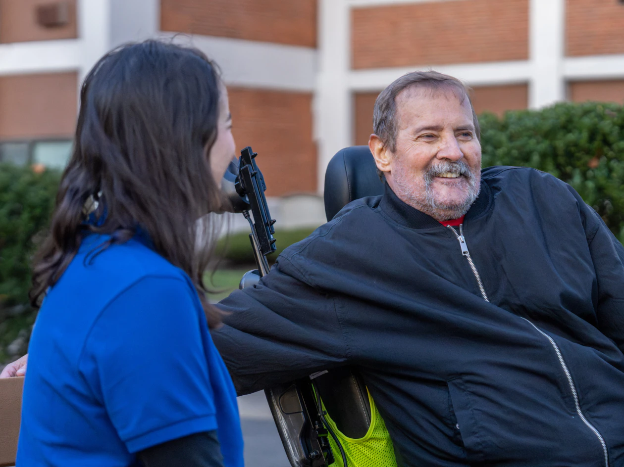 Image of a community health partner in a blue polo talking with a smiling man sitting in an electric wheelchair outdoors.