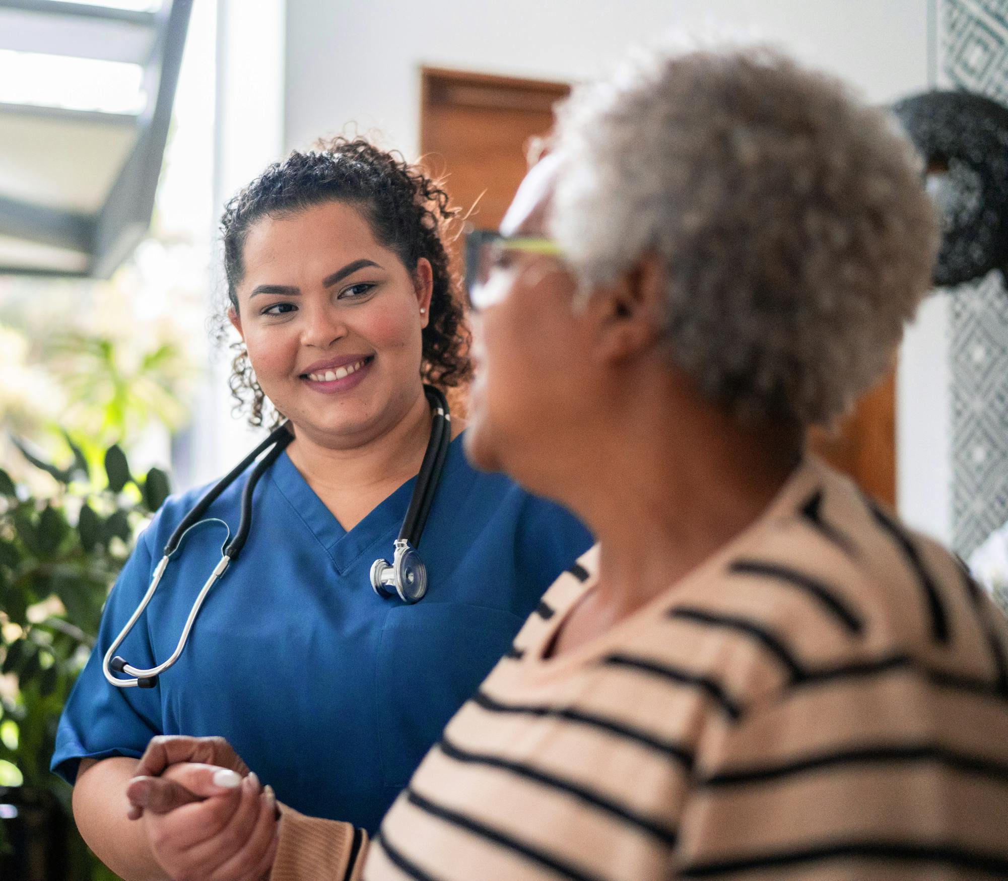 Healthcare worker wearing blue scrubs holds hand of person while smiling,