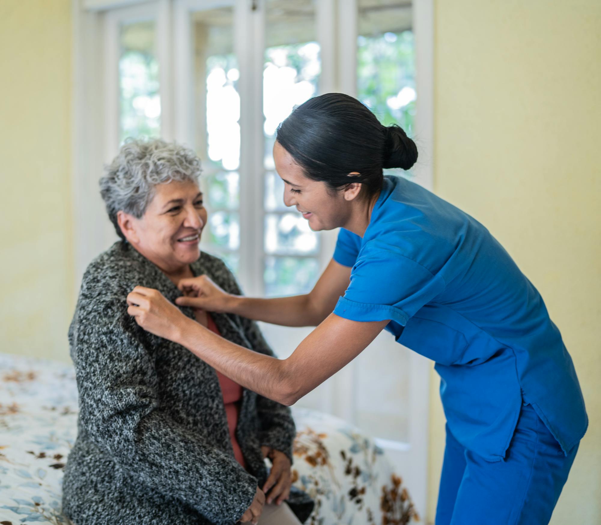 Healthcare worker wears blue scrubs while helping elderly woman at home.