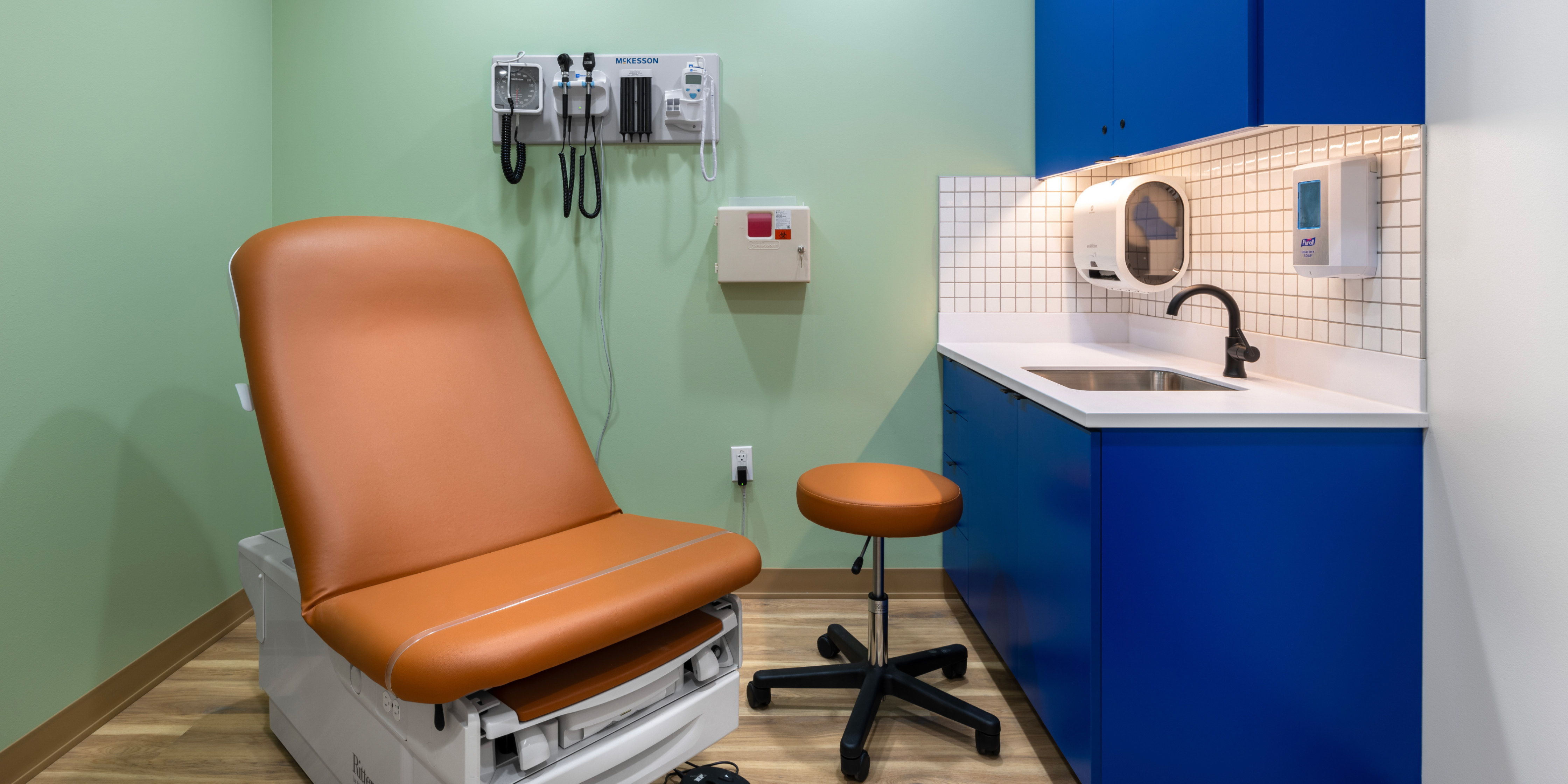 Cincinnati hub exam room with teal green walls, navy blue cabinets, and tan chairs.