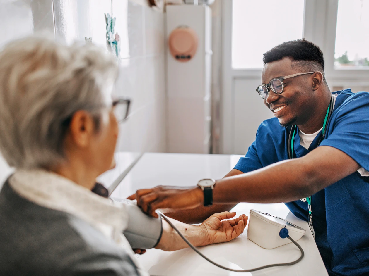 Image of a smiling male nurse in blue scrubs placing a blood pressure cuff on an older member's arm at a table.