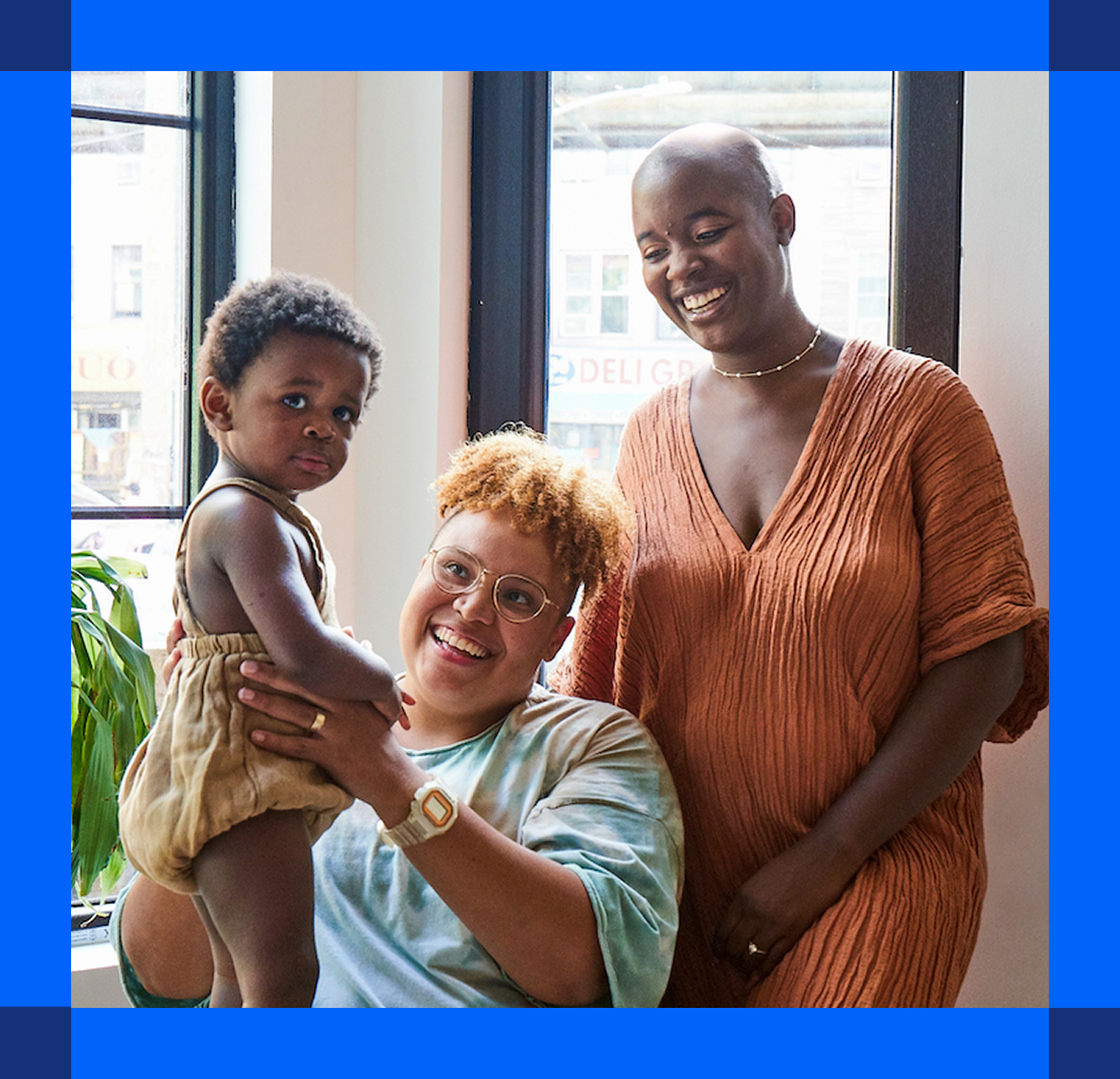 Person sits and holds their infant with their partner stands and smiles behind them.